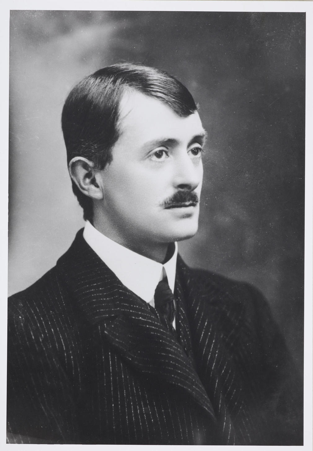 Photograph of a head and shoulders length portrait of the Poet Laureate and holder of the Order of Merit, John Masefield. He faces three-quarters right and wears a dark jacket, white shirt and tie.
