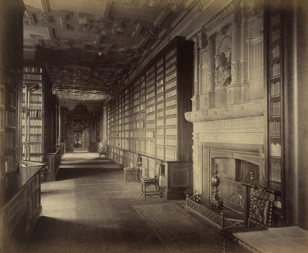 Queen Elizabeth's Gallery, in the Royal Library, WC. Shelves filled with books are either side of the walkway, chairs stand to the right. Window bays are on the left & a fireplace with carved niches and a bust above are (near) right.