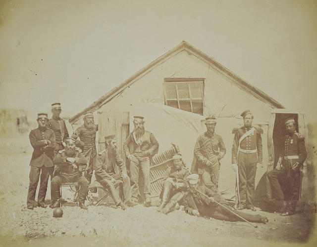 Group outside a hut, which is partly covered by canvas