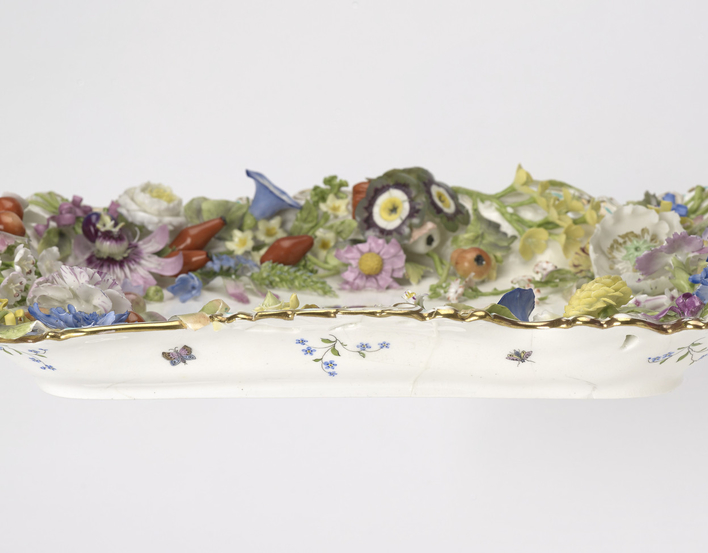 A rectangular porcelain tray painted in the centre with a crowned dated ribbon within a wreath. The border is encrusted with modelled coloured flowers (passion flowers, convolvulus, hips etc) and fruit. The bottom is painted with butterflies and floral sp